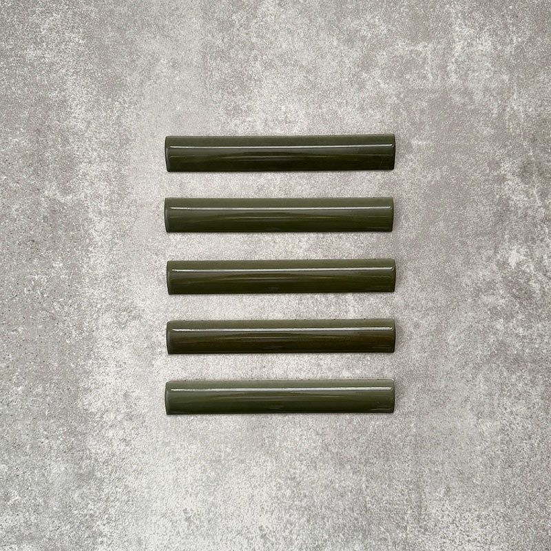 Flatlay von Zellige Botanisches Grün Bordürenfliese Keramik-Abschlussfliesen im marokkanischen Stil im Format 3 × 20 cm, in tiefen botanischen Grüntönen. Mit glatt abgerundetem Profil und glänzender Glasur auf einem strukturierten grauen Hintergrund arrangiert, um den handgefertigten Zellige-Effekt und das dekorative Abschlussprofil hervorzuheben.