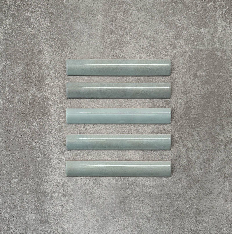 Flatlay mit Zellige Aquablau Bullnose-Keramikfliesen im marokkanischen Stil im Format 3 × 20 cm, die das abgerundete Kantenprofil, die glänzende blau-grüne Glasur und die feine tonale Variation vor neutralem Hintergrund zeigen.