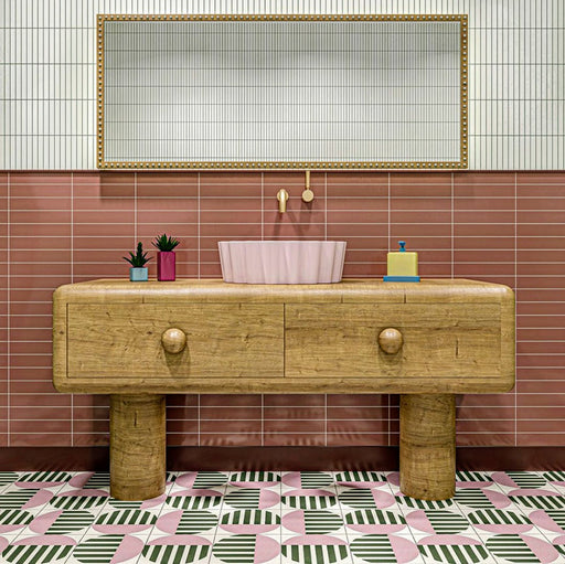 Badezimmerinterieur mit Abstrakter Serit Musterfliese im Format 20 × 20 cm, verlegt als markantes geometrisches Bodendesign. Das sich wiederholende Muster kombiniert sanfte Rosatöne, tiefes Grün und klares Weiß mit gestreiften Details und wird durch einen Waschtisch aus Holz sowie ein rosafarbenes Waschbecken ergänzt – für ein verspieltes und zugleich zeitgemäßes Badezimmerambiente.