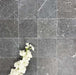 Flatlay mit Fayrouz Black Naturmarmorfliesen im Format 15,2 × 15,2 cm mit getrommelter Oberfläche. Tiefe Anthrazittöne und natürliche Maserungen werden durch weiche weiße Blumen stilvoll ergänzt und schaffen Kontrast sowie zusätzliche Textur.