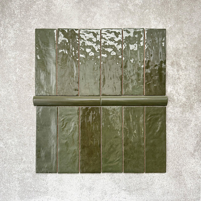 Flatlay mit Zellige Botanisches Grün Ziegel Keramikfliesen und passender Bullnose-Abschlussfliese: marokkanische Ziegel im Format 6,5 × 20 cm kombiniert mit einer 3 × 20 cm Bullnose-Kante in kräftigem botanischem Grün. Präsentiert auf neutralem, strukturiertem Hintergrund, um die glänzende Glasur, die natürliche Farbvariation und den handgefertigten Zellige-Effekt hervorzuheben.