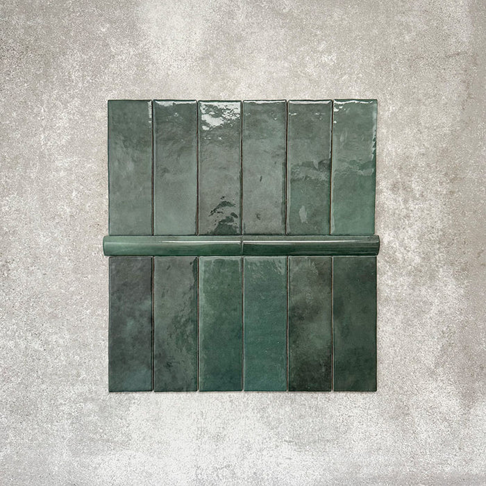Flatlay mit Zellige Königliches Grün Brick Keramikfliesen im marokkanischen Stil im Format 6,5 × 20 cm, kombiniert mit einer passenden Royal Green Bullnose-Abschlussfliese im Format 3 × 20 cm. Auf einem strukturierten grauen Hintergrund angeordnet, werden die glänzende Glasur, die feine tonale Variation und die handgefertigte Zellige-Optik in satten Grüntönen hervorgehoben.