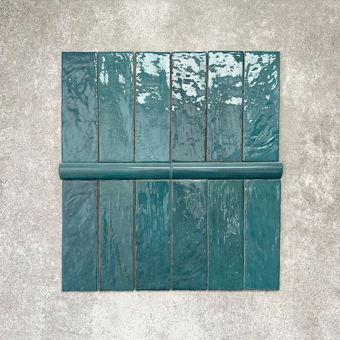 Flatlay mit Zellige Pfauengrün Keramikfliesen im marokkanischen Stil, bestehend aus 6,5 × 20 cm Ziegelfliesen kombiniert mit einer passenden 3 × 20 cm Bullnose-Abschlussfliese. Hervorgehoben werden das abgerundete Kantenprofil, die glänzende tief blau-grüne Glasur und die natürliche Farbvariation auf neutralem Hintergrund.