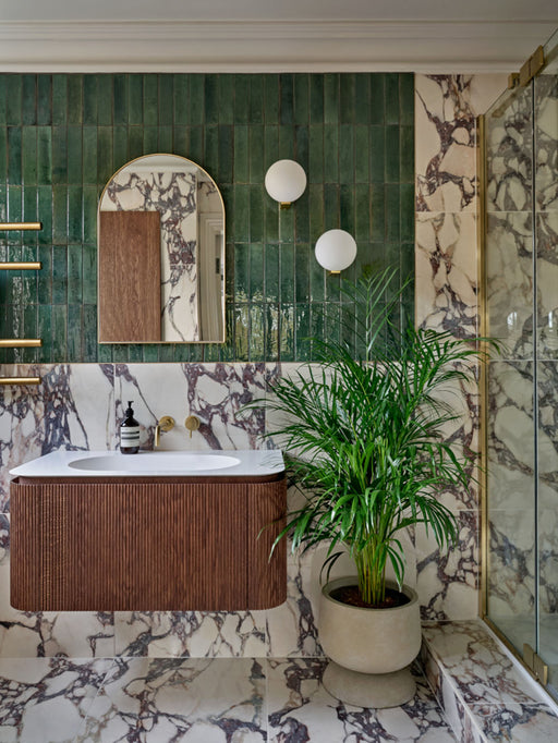 Badezimmerumgebung mit glänzenden Elazar Olivgrün Ziegelfliesen im Format 6 × 25 cm, kombiniert mit Fayrouz Viola 30,5 × 61 cm Naturmarmorfliesen. Die satten Grüntöne treffen auf markante bordeauxrote Maserung und werden durch warme Holzstrukturen sowie Messingarmaturen ergänzt – für ein raffiniertes, vielschichtiges Gesamtbild.