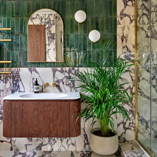 Badezimmerumgebung mit glänzenden Elazar Olivgrün Ziegelfliesen im Format 6 × 25 cm, kombiniert mit Fayrouz Viola 30,5 × 61 cm Naturmarmorfliesen. Die satten Grüntöne treffen auf markante bordeauxrote Maserung und werden durch warme Holzstrukturen sowie Messingarmaturen ergänzt – für ein raffiniertes, vielschichtiges Gesamtbild.