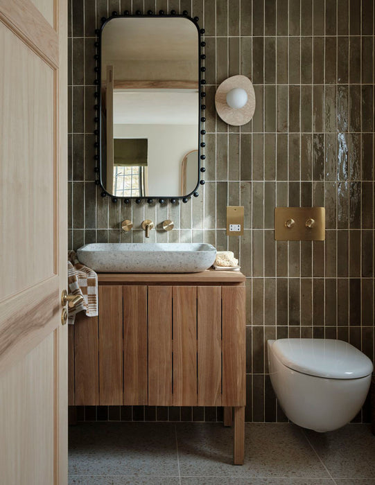 Badezimmer mit Elazar Salbeigrün glänzender Ziegel-Fliese im Format 6x25 cm, vertikal verlegt hinter Holzwaschtisch und Spiegel, kombiniert mit Messingarmaturen für eine warme, moderne Wandgestaltung.