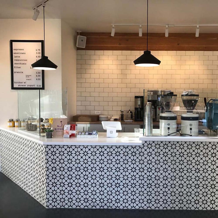 Café-Theke mit Agadir Schwarz & Weiß Fliesen im marokkanischen Stil im Format 20 × 20 cm, eingesetzt als dekorative Front. Kombiniert mit weißen Metro-Wandfliesen, warmen Holzakzenten und moderner schwarzer Beleuchtung.