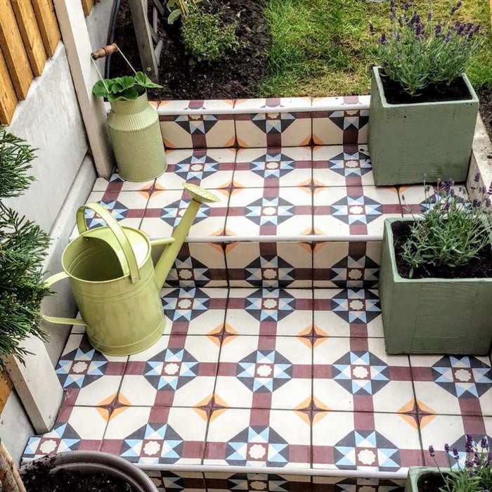 Gartentreppe mit Berlin Kreuz Bodenfliesen im Format 20 × 20 cm im viktorianisch-marokkanisch inspirierten Stil. Inszeniert mit Topfpflanzen und einer Gießkanne, zeigt sich ein dekoratives Outdoor-Fliesendesign in sanften Blau-, Burgunder- und Cremetönen.