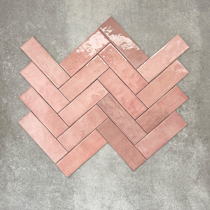 Flatlay von Zellige Rosa Brick Keramikfliesen im marokkanischen Stil im Format 6,5 × 20 cm, in sanften Blush-, Altrosa- und Terrakotta-Pinktönen mit feiner tonaler Variation und leicht unregelmäßigen Kanten. Im Fischgrätmuster auf strukturiertem grauem Hintergrund arrangiert, um die handgefertigte Zellige-Glasur hervorzuheben.
