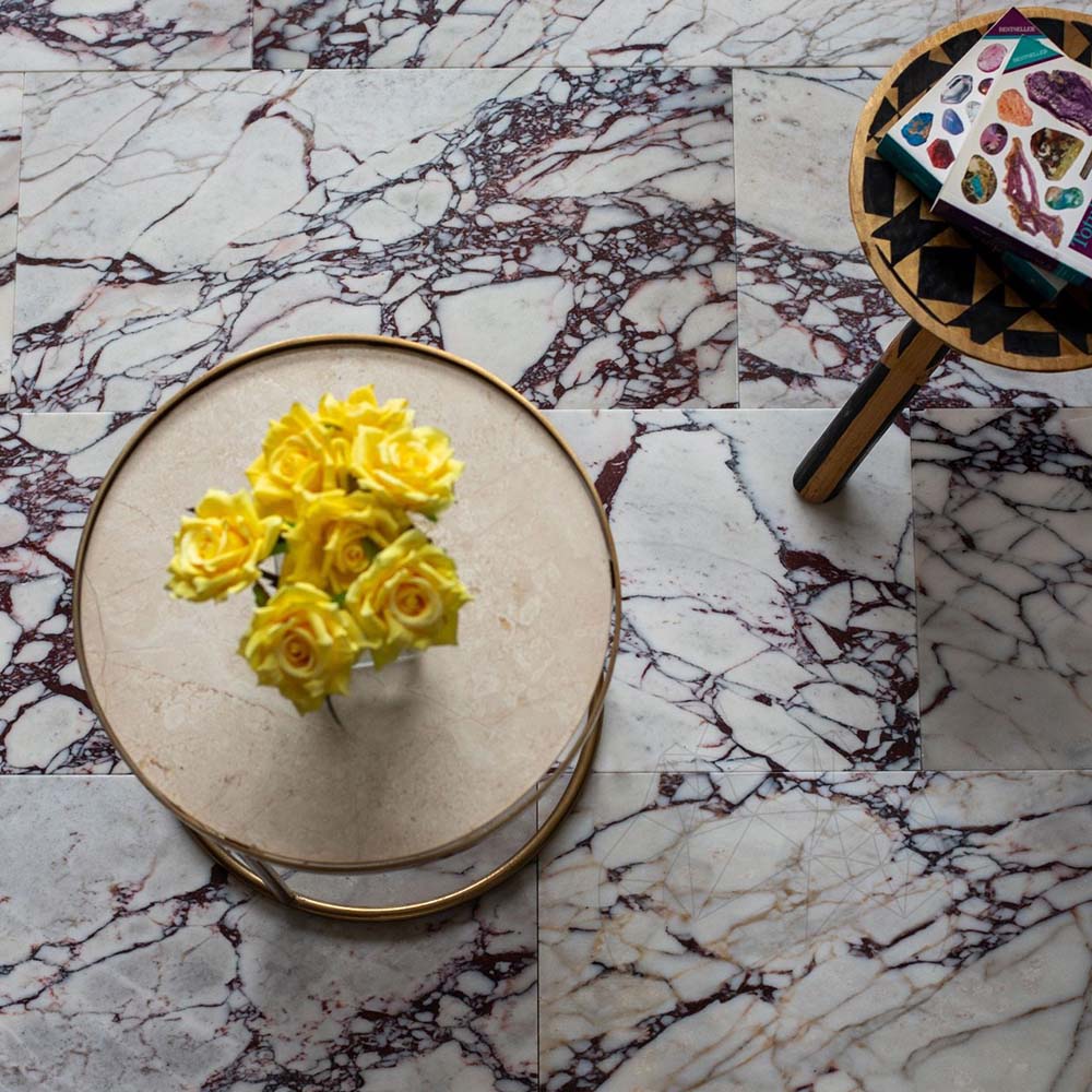 Innenraum mit Fayrouz Viola 30,5 × 61 cm Naturmarmorfliesen mit geschliffener Oberfläche, im versetzten Verband verlegt, mit weißer Basis und markanter bordeauxroter Maserung, kombiniert mit einem runden Tisch und dekorativen Accessoires.