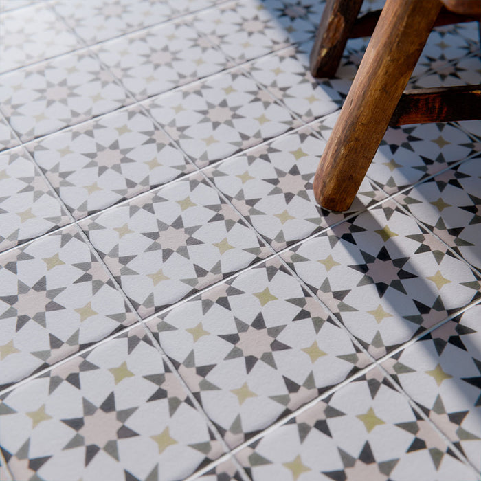 Marketingmotiv mit Agadir Beige Fliesen im marokkanischen Stil im Format 20 × 20 cm aus Feinsteinzeug, verlegt als Bodenbelag mit einem sanften, neutralen geometrischen Sternmuster in dezenten Grau- und warmen Beigetönen. Mit einem Holzstuhl in Szene gesetzt, um eine wohnliche, authentische Atmosphäre zu vermitteln.