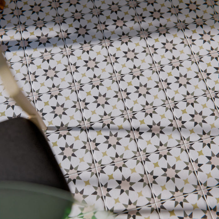 Marketingmotiv mit Agadir Beige Fliesen im marokkanischen Stil im Format 20 × 20 cm aus Feinsteinzeug, verlegt als Bodenbelag mit einem sanften, neutralen geometrischen Sternmuster in dezenten Grau- und warmen Beigetönen. Mit einem Holzstuhl in Szene gesetzt, um eine wohnliche, authentische Atmosphäre zu vermitteln.