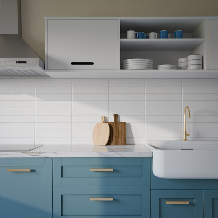 Moderne Küche mit Bora Bora Ziegel Weißer Sand Keramik-Wandfliesen im schlanken Riemchenformat 6,5 × 32,8 cm mit Fluxglasur, horizontal als Rückwand hinter blauen Schrankfronten verlegt. Kombiniert mit weißen Arbeitsplatten, offenen Regalen und Armaturen in Messing entsteht ein klarer, zeitgemäßer Look.