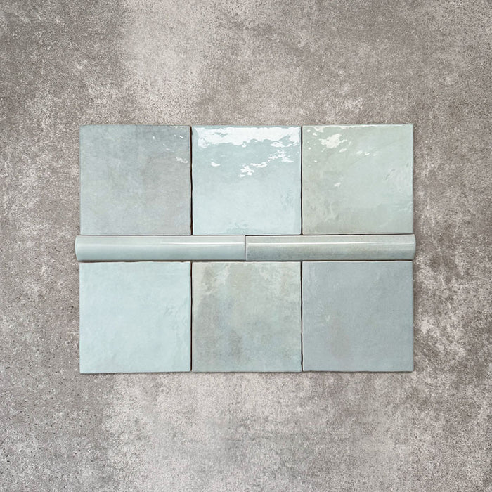 Flatlay mit Zellige Aquablau Keramikfliesen im marokkanischen Stil, bestehend aus 13,2 × 13,2 cm Quadratfliesen in Kombination mit einer 3 × 20 cm Bullnose-Abschlussfliese. Gezeigt werden das abgerundete Kantenprofil, die glänzende blau-grüne Glasur und die feine tonale Variation vor neutralem Hintergrund.