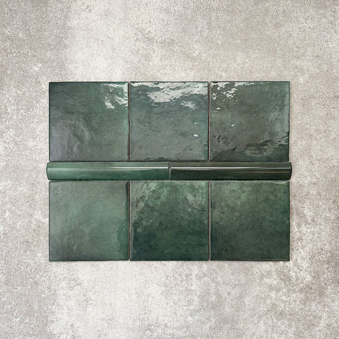 Flatlay mit Zellige Königliches Grün Quadrat Keramikfliesen im marokkanischen Stil im Format 13,2 × 13,2 cm, kombiniert mit einer passenden Royal Green Bullnose-Abschlussfliese im Format 3 × 20 cm. Satter Grünton, natürliche Glasurvariation und leicht unregelmäßige Kanten werden auf einem strukturierten grauen Hintergrund gezeigt und unterstreichen den handgefertigten Zellige-Effekt.