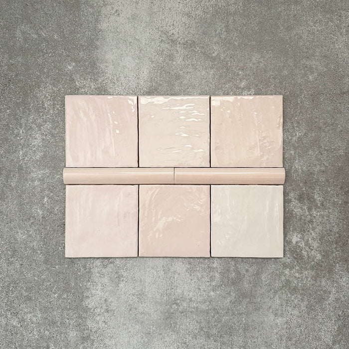 Flatlay mit Zellige Rosa Nebel Quadrat Keramikfliesen im marokkanischen Stil im Format 13,2 × 13,2 cm, kombiniert mit einer passenden Pink Mist Bullnose-Abschlussfliese im Format 3 × 20 cm. Sanfte, helle Rosétöne, natürliche Glasurvariation und leicht unregelmäßige Kanten werden auf einem strukturierten grauen Hintergrund gezeigt und unterstreichen die handgefertigte Zellige-Optik.