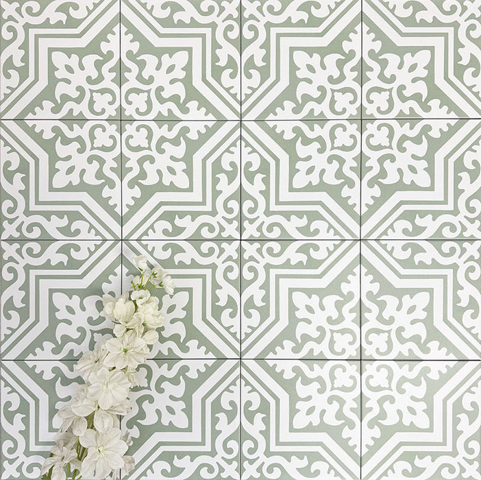 Flatlay mit Berlin Trondite Grün Fliesen im Format 20 × 20 cm im viktorianisch-marokkanisch inspirierten Stil, verlegt in einem sich wiederholenden Muster. Feine weiße Ornamente auf einem salbeigrünen Hintergrund werden durch dekorative weiße Blumen ergänzt.