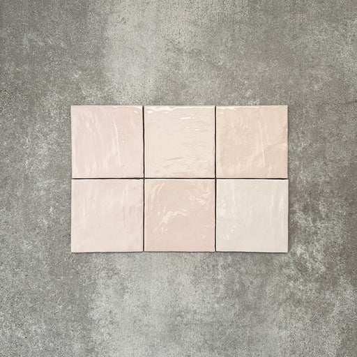 Flatlay von Zellige Rosa Nebel Quadrat Keramikfliesen im marokkanischen Stil im Format 13,2 × 13,2 cm, in zarten Rosa- und Blushtönen. Im schlichten Raster auf strukturiertem grauem Hintergrund arrangiert, um die glänzende Glasur, die feine tonale Variation und die handgefertigte Zellige-Optik hervorzuheben.