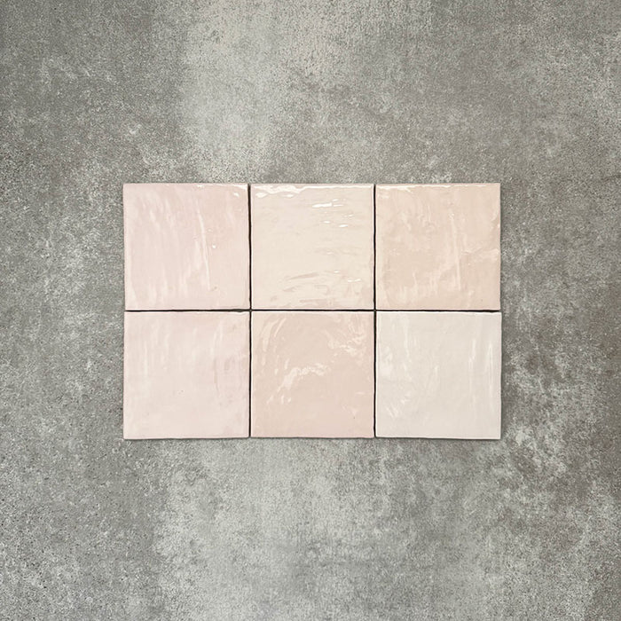 Flatlay von Zellige Rosa Nebel Quadrat Keramikfliesen im marokkanischen Stil im Format 13,2 × 13,2 cm, in zarten Rosa- und Blushtönen. Im schlichten Raster auf strukturiertem grauem Hintergrund arrangiert, um die glänzende Glasur, die feine tonale Variation und die handgefertigte Zellige-Optik hervorzuheben.