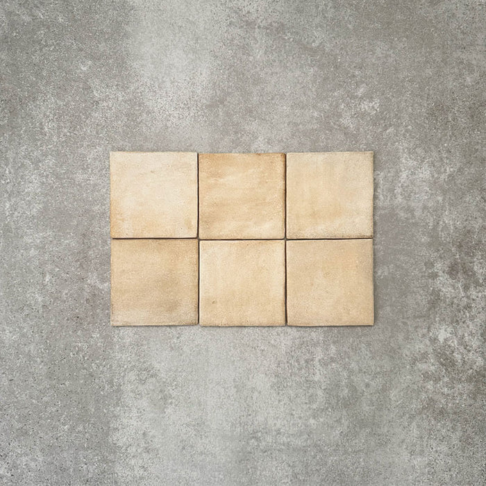 Flatlay von Zellige Sandbeige Matt Quadrat Keramikfliesen im marokkanischen Stil im Format 10 × 10 cm, in warmen Beige- und Sandnuancen. Dezente Farbvariation und sanft unregelmäßige Kanten sind in einem schlichten Raster auf strukturiertem grauem Hintergrund arrangiert, um die handwerkliche Zellige-Optik hervorzuheben.