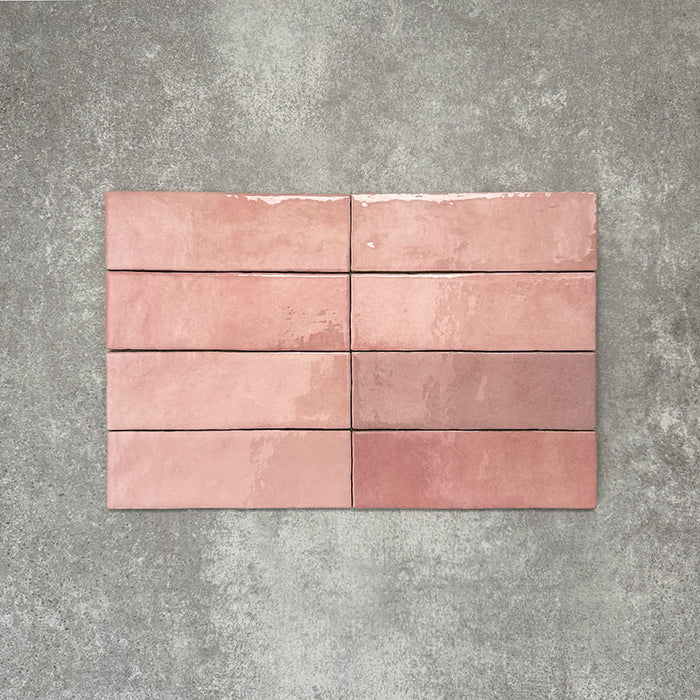 Flatlay von Zellige Rosa Brick Keramikfliesen im marokkanischen Stil im Format 6,5 × 20 cm, in sanften Blush-, Altrosa- und Terrakotta-Pinktönen mit feiner tonaler Variation und leicht unregelmäßigen Kanten. Vertikal im Ziegelverband auf strukturiertem grauem Hintergrund arrangiert, um die handwerkliche Zellige-Optik zu zeigen.