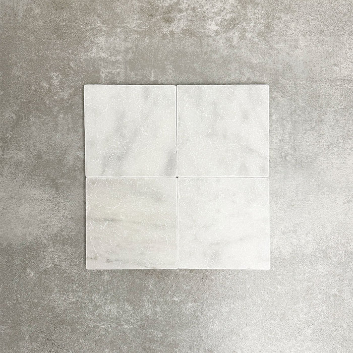 Vier Fayrouz White 15,2 × 15,2 cm Naturmarmorfliesen mit getrommelter Oberfläche nebeneinander angeordnet, die sanften Weißtöne, feine Maserung und natürliche Variation vor einem neutralen, strukturierten Hintergrund zeigen.