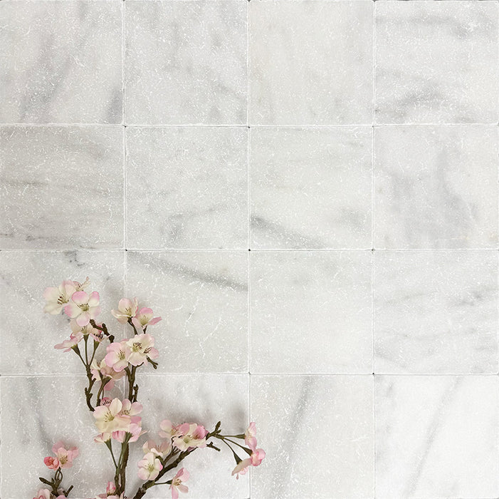 Flatlay mit Fayrouz White 15,2 × 15,2 cm Naturmarmorfliesen mit getrommelter Oberfläche in sanften Weißtönen mit dezenter natürlicher Maserung, kombiniert mit zarten rosa Blütenzweigen für eine ruhige, elegante Ausstrahlung.